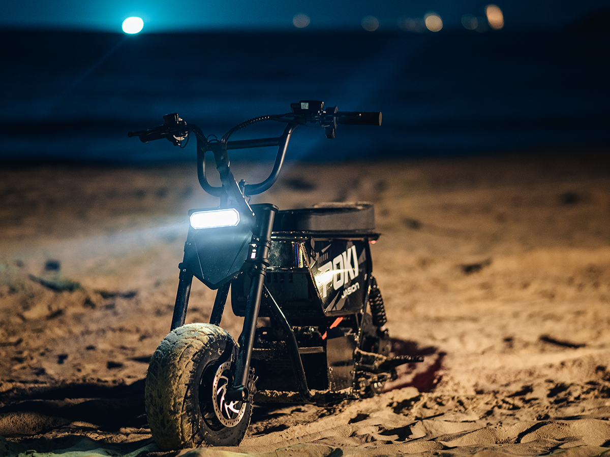 Black Jasion Poki Electric Mini Bike parked on sand at night with its headlight illuminated