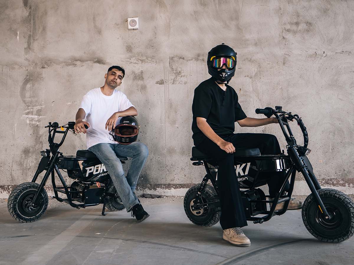 Two riders seated on black Jasion Poki electric mini bikes beside a concrete wall